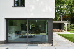 Přechod mezi zahradou a domem tvoří betonové desky a rýnský štěrkopísek. Tmavé rámové profily prosklených ploch kontrastují se světlými povrchy.