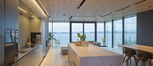 Obývací pokoj, kuchyně i jídelna sloučené do rodinného pokoje. V prosvětleném, jednoduchém architektonickém řešení interiéru hraje ústřední roli panorama krajiny (fasádní systém Schüco FWS 50.HI v kombinaci s posuvným systémem se zdvihem Schüco ASS 70.HI), foto Sindre Ellingsen 