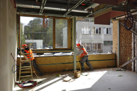 Na podélné plnostěnné vazníky se připevnily kotvy pro zavěšení lehkého dřevěného obvodového pláště o tloušťce 24 cm. Výroba prefabrikátů probíhala za pomoci CNC technologií, takže bylo dosaženo vysoké přesnosti a kvality. Hlavní předností systému ENVILOP je nicméně rychlost realizace – obvodové panely o rozměrech 3 x 3,6 m na stavbu přijížděly zkompletované s okny a fasádami. Při osazování byly panely zavěšeny na kotvy pomocí rektifikačních šroubů. Ocelové kotvy pro obklad se do zdiva připevnily přes podložku termostop.