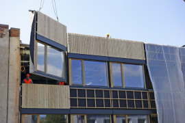 Na podélné plnostěnné vazníky se připevnily kotvy pro zavěšení lehkého dřevěného obvodového pláště o tloušťce 24 cm. Výroba prefabrikátů probíhala za pomoci CNC technologií, takže bylo dosaženo vysoké přesnosti a kvality. Hlavní předností systému ENVILOP je nicméně rychlost realizace – obvodové panely o rozměrech 3 x 3,6 m na stavbu přijížděly zkompletované s okny a fasádami. Při osazování byly panely zavěšeny na kotvy pomocí rektifikačních šroubů. Ocelové kotvy pro obklad se do zdiva připevnily přes podložku termostop.